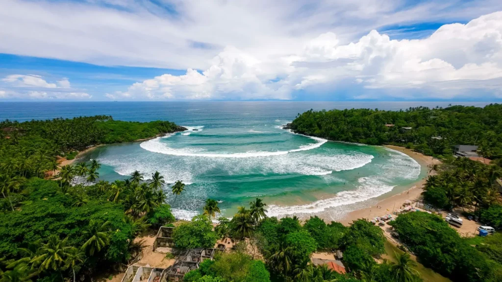 A breathtaking aerial drone shot of the perfect horseshoe-shaped bay at Hiriketiya beach, showing turquoise waters fringed by dense coconut palms